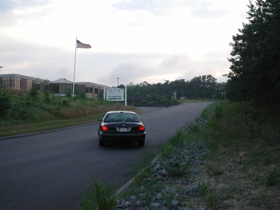 Dark sedan driving away from camera on a road, with a building and American flag in background