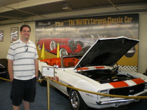 1998 Ford Crown Victoria - man poses next to a white Chevrolet Camaro SS convertible with orange stripes and open hood at a classic car museum.