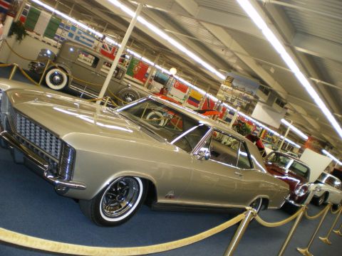 1998 Ford Crown Victoria - silver mid-1960s Buick Riviera coupe with whitewall tires displayed behind velvet ropes at a classic car museum.