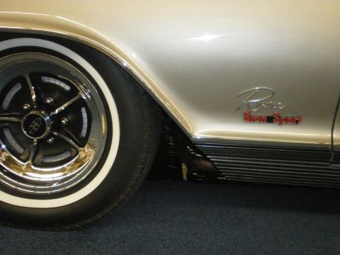 1998 Ford Crown Victoria - Detail of a gold Buick Riviera Gran Sport showing its chrome five-spoke mag wheel, whitewall tire and red script badge.