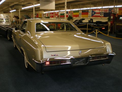 1998 Ford Crown Victoria - Gold Buick Riviera hardtop coupe shown from the rear in a museum hall hung with international flags.