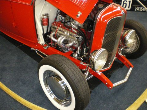 1998 Ford Crown Victoria - Red hot rod roadster showing its polished multi-carburetor engine, chrome headlight and wide whitewall front tire.