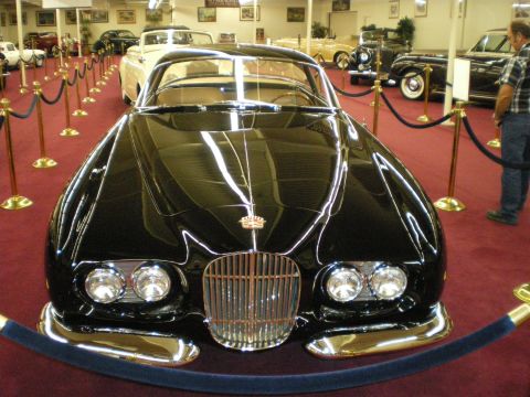 1998 Ford Crown Victoria - Black streamlined custom coupe with a tall chrome grille and quad headlights displayed in a red-carpeted museum.