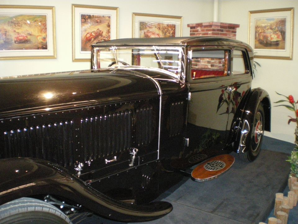 Side rear view of a dark prewar coupe with louvered hood and side-mounted spare on a stage