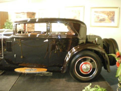 1998 Ford Crown Victoria - Dark prewar sedan displayed in profile with side-mounted spare wheels and red-accented wheels on a museum stage.