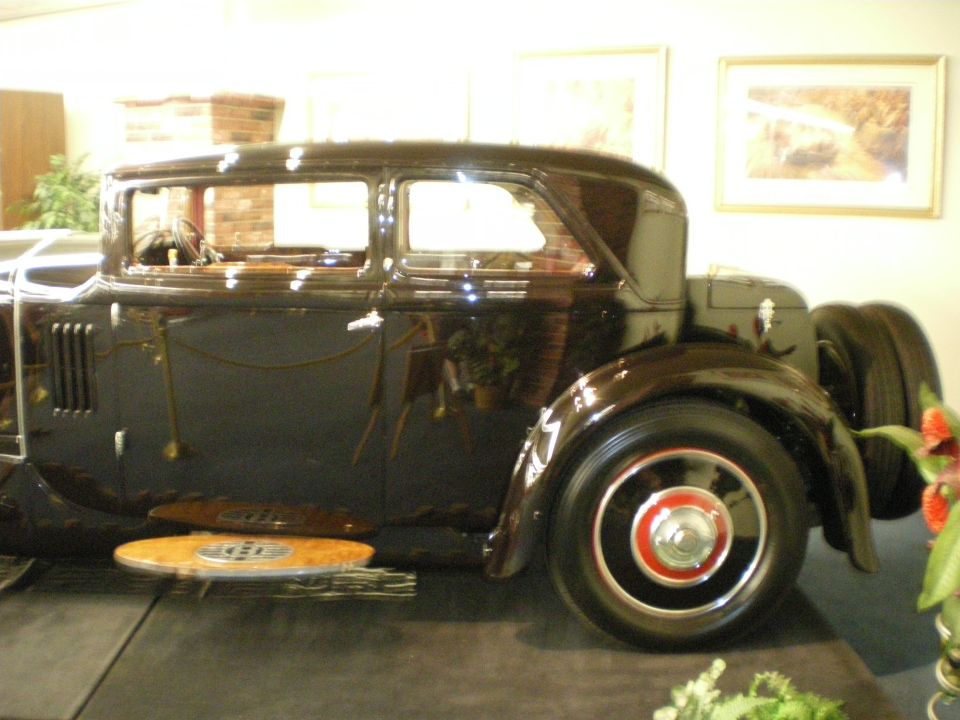 Side view of a dark prewar sedan with side-mounted spares and red-accented wheels on a stage
