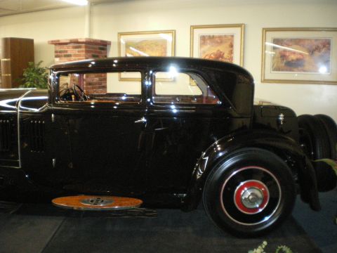 1998 Ford Crown Victoria - Black prewar coupe shown in profile with a louvered hood and red-pinstriped wheels on a carpeted display stage.