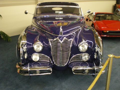 1998 Ford Crown Victoria - Front of a deep-purple custom classic coupe with prominent vertical chrome grille, beside a red Ferrari in a showroom.