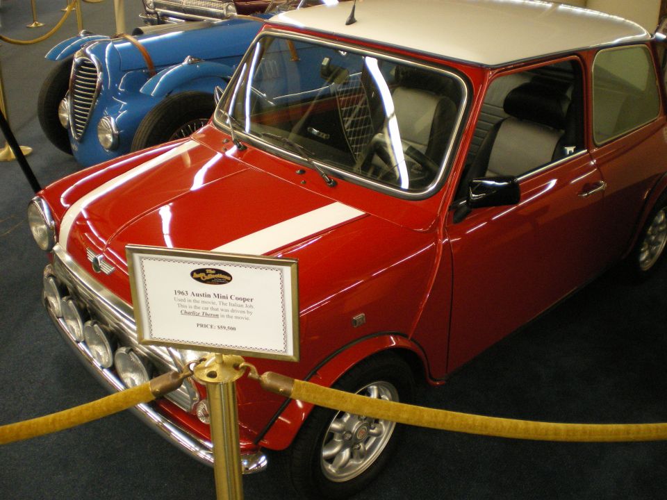 Red Mini Cooper with white roof on display behind a printed placard, blue car behind