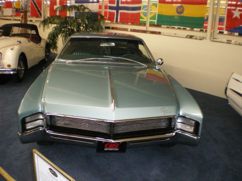 1998 Ford Crown Victoria - Front view of a light-teal 1960s personal-luxury coupe with hidden-headlamp full-width grille below a row of flags.