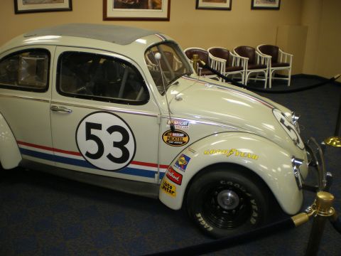 1998 Ford Crown Victoria - cream VW Beetle in Herbie livery with number 53, red-and-blue stripes and racing decals displayed indoors.
