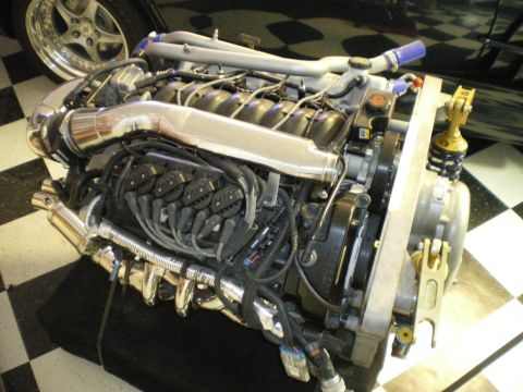 1998 Ford Crown Victoria - bare modern engine with intake manifold, headers and wiring harness sitting on a checkered-floor display stand.