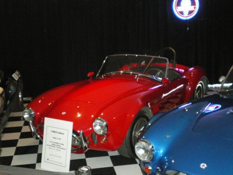 1998 Ford Crown Victoria - red Cobra-style roadster beside a blue roadster on a checkered floor under a lit emblem, with an info placard.