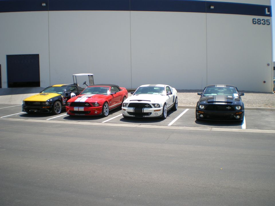 Four Ford Shelby Mustangs in yellow, red, white and black parked outside a warehouse
