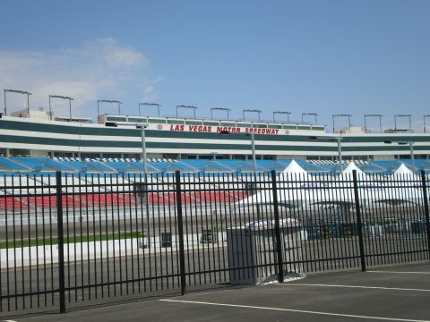 1998 Ford Crown Victoria - green-and-white grandstands of Las Vegas Motor Speedway viewed through a black iron perimeter fence.