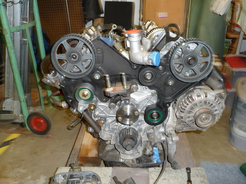 V6 engine on wooden blocks in a garage with timing belt removed, exposing cam pulleys, water pump and alternator.
