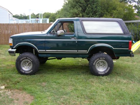 1993 Ford Bronco - This lifted 1993 Ford Bronco features a dark green paint job and custom wheels, seen parked on a grassy lawn.