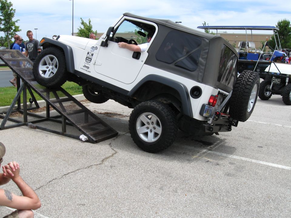 White Jeep Wrangler with a soft top on a metal ramp, front wheels elevated, with people watching