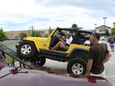 1994 Ford Bronco - A yellow Jeep Wrangler is driven up a ramp at a car show, with onlookers and other vehicles present.