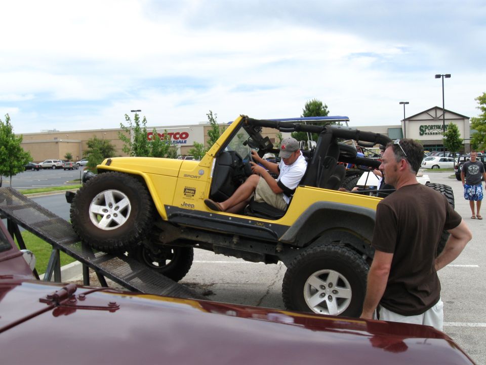 Yellow Jeep Wrangler on a ramp with a man driving, another man watching, and a maroon vehicle in the foreground