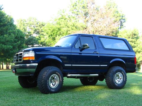 1967 Ford Mustang - A dark blue Ford Bronco is shown from the side, parked on a grassy lawn with trees in the background.