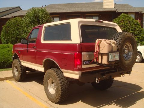 1992 Ford Bronco - Three-quarter rear view of a lifted red and cream two-tone Ford Bronco with a white removable hardtop, parked at a brick apartment complex…