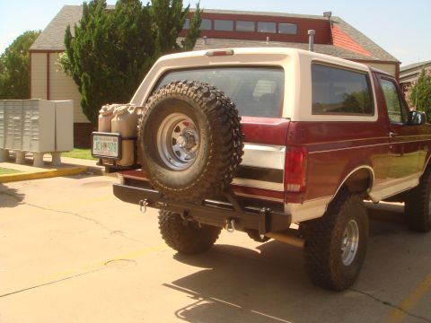 1992 Ford Bronco - Three-quarter rear view of a lifted two-tone maroon and tan full-size Ford Bronco parked on a concrete lot, wearing a black tubular rear bu…