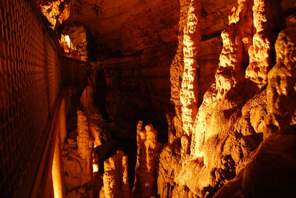 Inside a cave with stalactites and stalagmites, illuminated by orange lights. A metal walkway is visible on the left