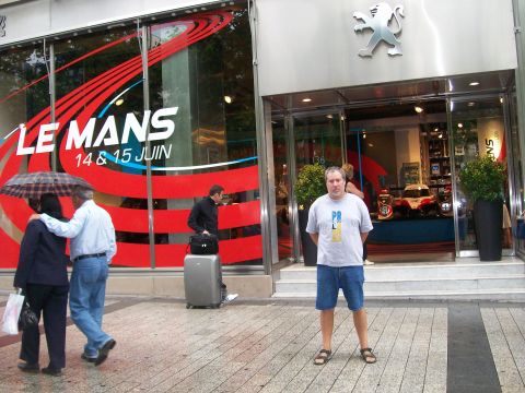 2008 Citroen Xantia - A man poses outside a Peugeot dealership a Le Mans advertisement, while others walk by under an umbrella.