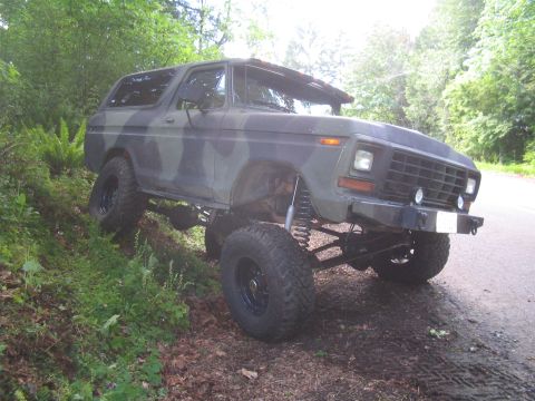1979 Ford Bronco - This heavily modified 1978 Ford Bronco features an extreme lift kit and camouflage paint, parked on the side of a wooded road.