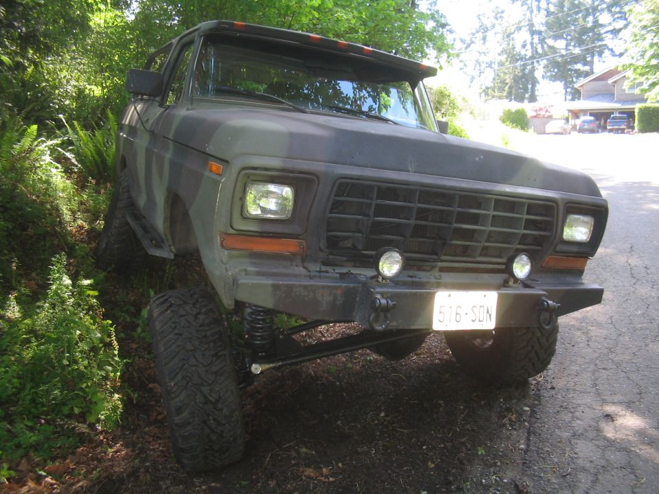 Camouflaged Ford Bronco lifted with big tires, parked on a dirt path next to some ferns