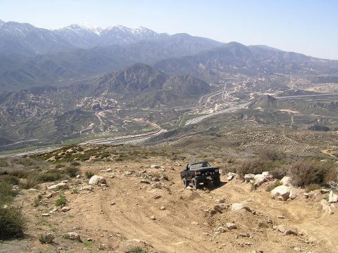 1990 Ford Bronco - A black 1990 Ford Bronco navigates a , rocky terrain with a vast mountain range and highway visible in the distance.