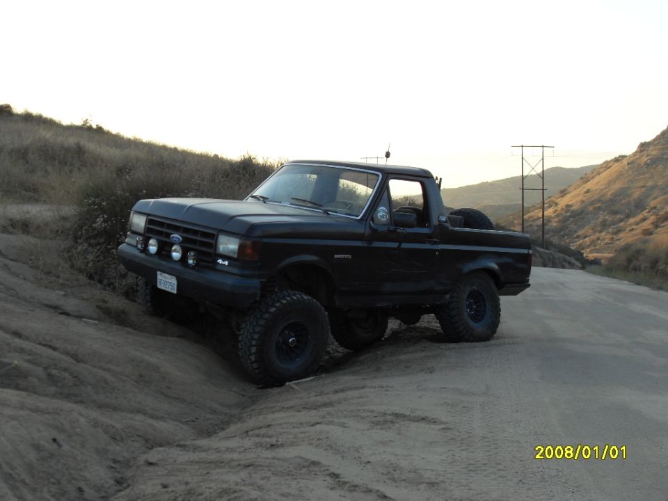 1990 Ford Bronco with off-road tires climbing a dirt hill, spare tire mounted on rear