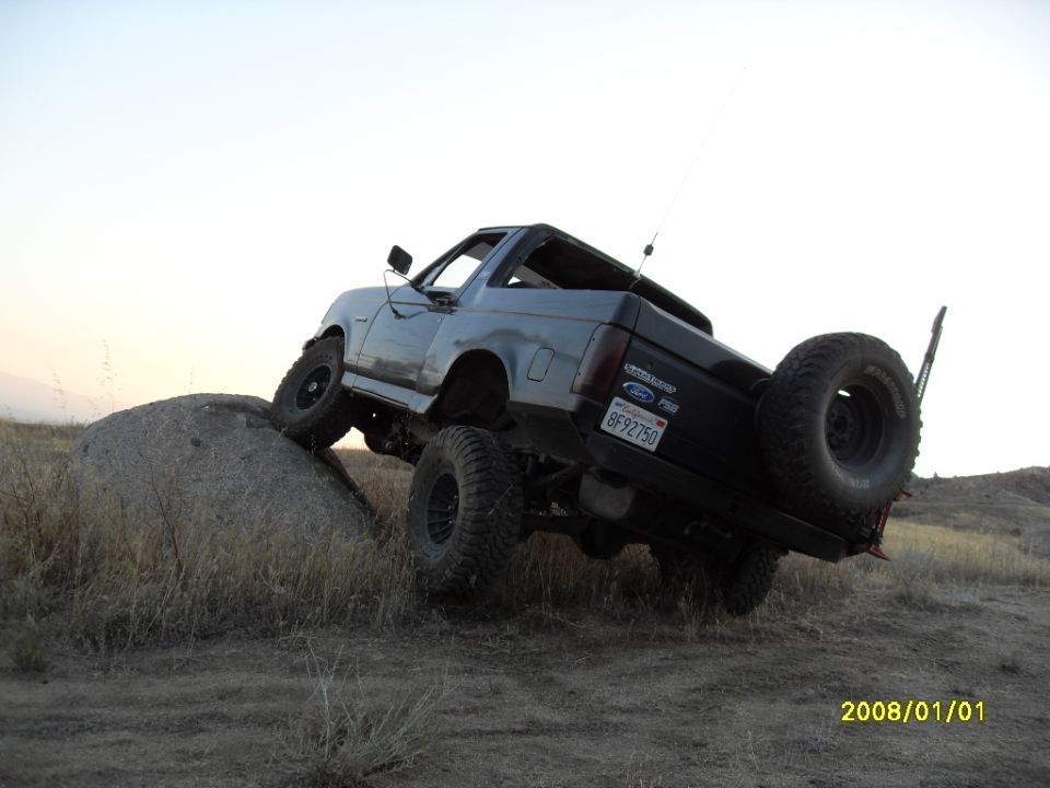 Gray 1990 Ford Bronco off-roading on a rock, rear-mounted spare tire visible