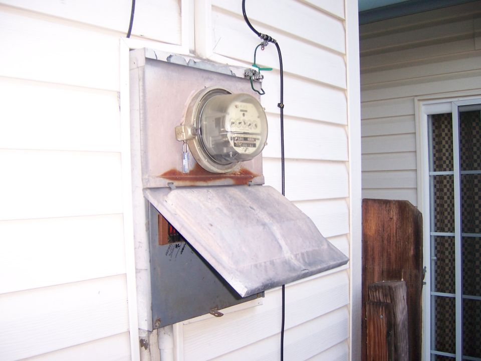 Outdoor electrical meter box mounted on a white sided house. The meter is visible behind glass, and a metal flap is open below it