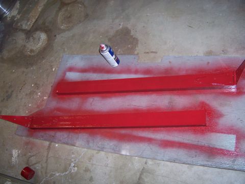 1991 Ford Bronco - Red metal bars are being spray painted on a plywood sheet on a concrete floor, with a can of spray paint nearby.