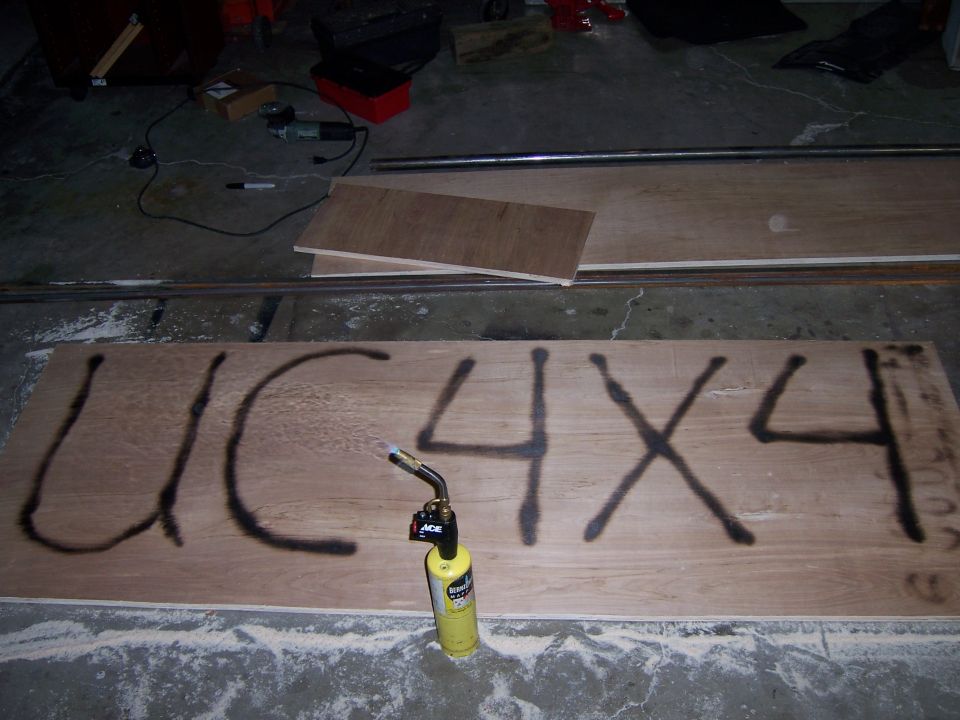 Plywood sheet with black spray-painted text "UC 4X4" and a yellow propane torch
