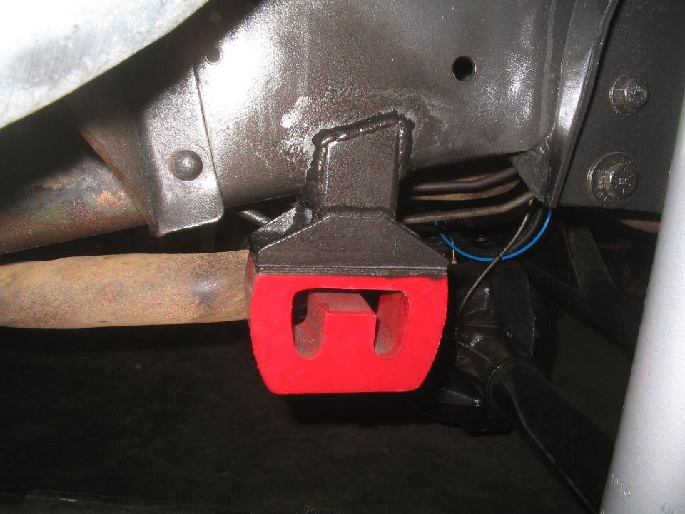 Close-up of a red polyurethane bushing mounted to a vehicle's frame, near a rusty exhaust pipe