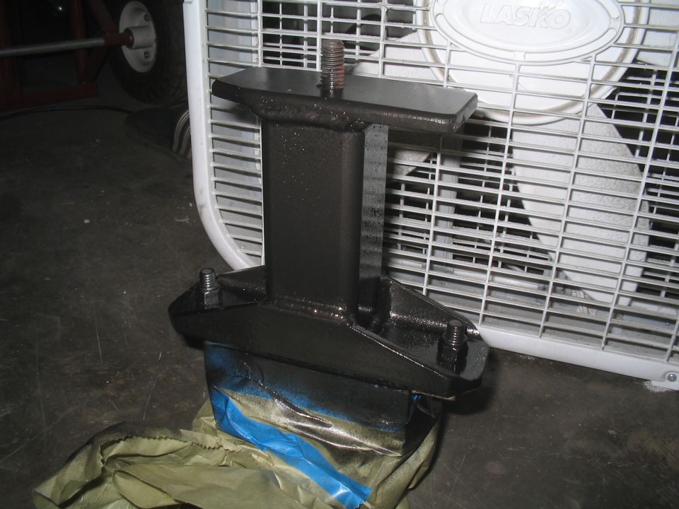 Black metal bracket with bolts and threaded rod, partially covered in blue tape, in front of a white box fan