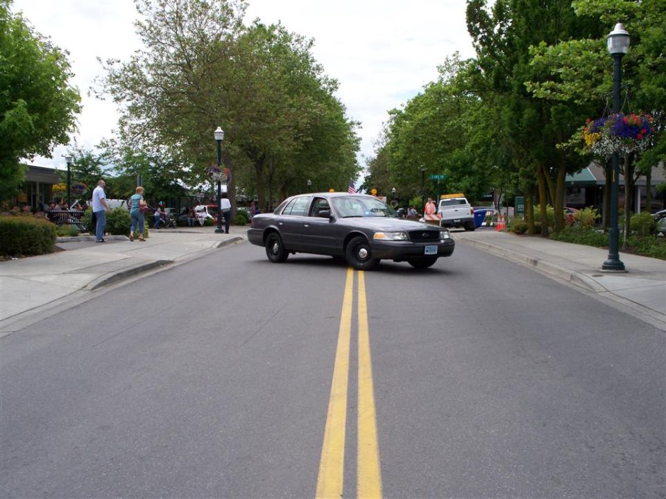 Dark gray Ford Crown Victoria sedan crossing a tree-lined town street