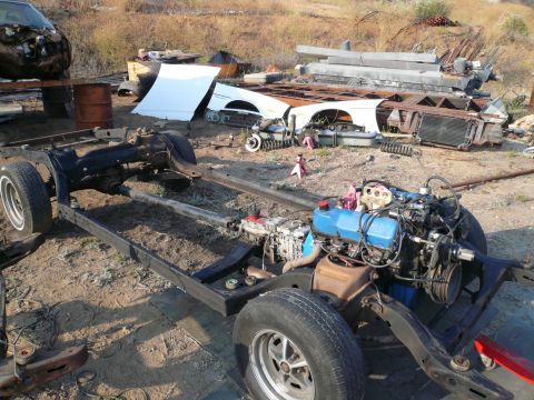 1975 Ford Ranchero - Disassembled vehicle frame and engine in a junkyard setting with scattered car parts.