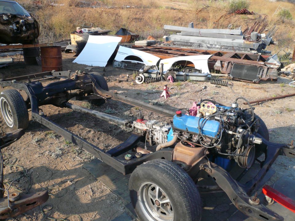 Car frame with engine, wheels, and various disassembled parts scattered around. A dark vehicle is partially visible on a lift in the background