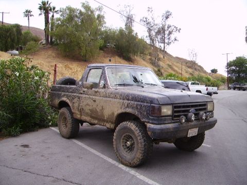 1990 Ford Bronco - Mud-covered 1990 Ford Bronco parked in a lot, with other vehicles visible behind it.