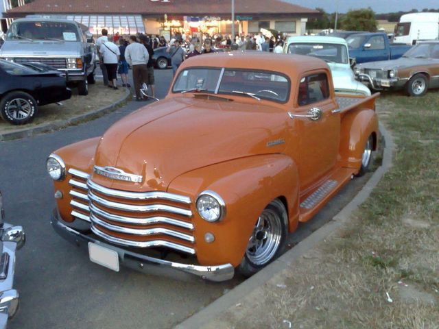 Orange Chevrolet pickup truck with chrome grille and custom wheels parked on a curb