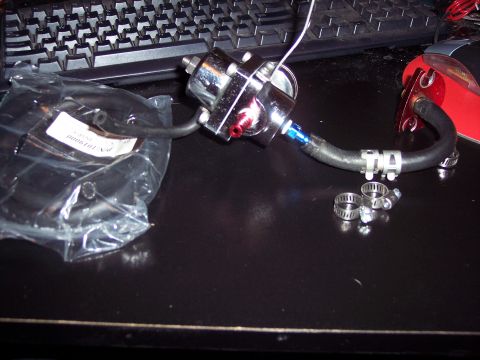 1995 Ford Probe - A chrome fuel pressure regulator with black hose and red fittings is shown on a dark surface next to a keyboard.
