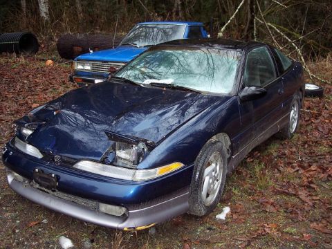1995 Ford Probe - A dark blue coupe with its pop-up headlights partially open sits in a wooded area next to a blue pickup truck.