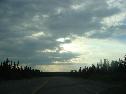 2001 Chevrolet Corvette - A desolate road winds through a forest under a dramatic, cloudy sky, suggesting a journey through remote wilderness.