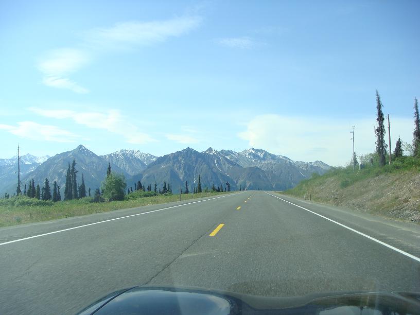 View from a car dashboard looking out at a highway stretching towards snow-capped mountains under a blue sky