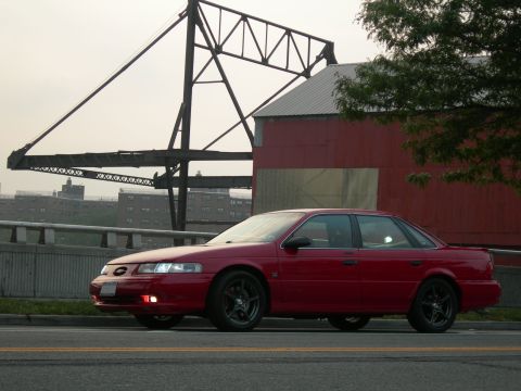 1992 Ford Taurus - A red 1992 Ford Taurus sedan is captured from a three-quarter front view, with a red barn and industrial background.