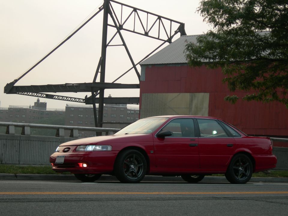 Red Ford Taurus SHO sedan on black wheels parked by a red industrial building